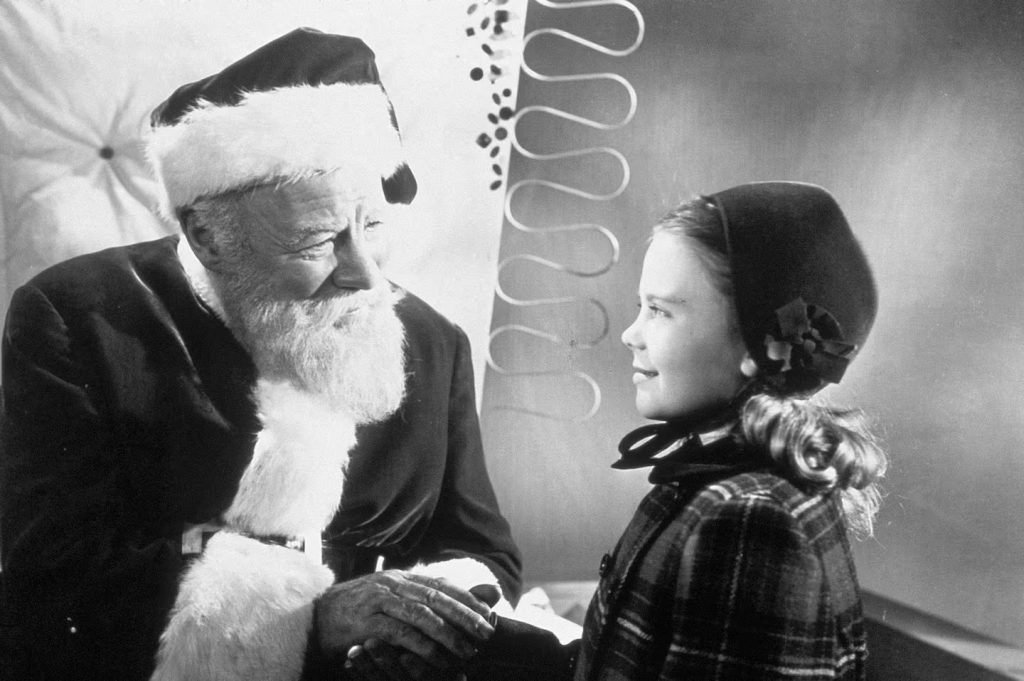 A black and white still from the 1947 movie Miracle 34th Street. An older man with a white beard and wearing a Santa costume smiles at a young girl with curly hair wearing a black hat and a plaid coat.