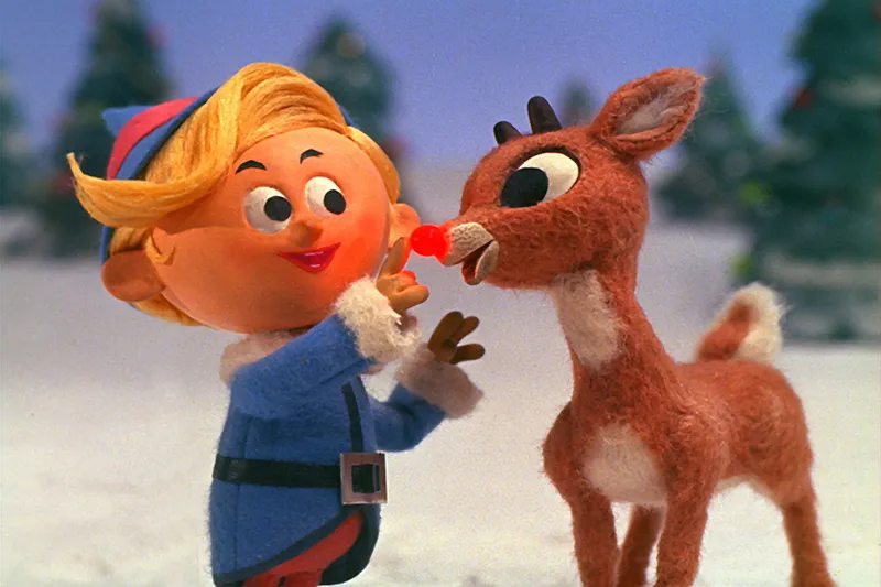 A still from the stop motion film Rudolph the Red-Nosed Reindeer. A winter landscape with decorated pine trees in the background. A young reindeer with stubby antlers and a glowing red nose looks at an elf with coiffed blonde hair wearing a blue and pink elf costume trimmed in white fur. The elf reaches up to touch the reindeer's nose.