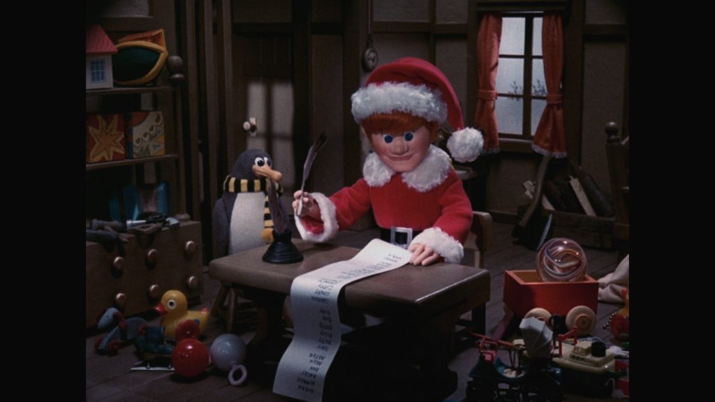 A still from the stop motion film Santa Claus Is Comin' to Town. A young man with pale skin and red hair wears a red Santa Claus costume. He writes with a quill pen on a long sheet of white paper. Beside him sits a penguin wearing a yellow and black striped scarf. They are in a wooden room surrounded by toys.