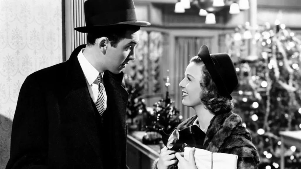 A black and white still from the movie The Shop Around the Corner. A man in a dark hat and coat looks down at a smiling woman wearing a black hat and fur coat clutching a package. Behind them is a room full of decorated Christmas trees.