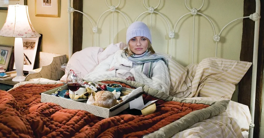 A still from the movie The Holiday. A young woman with pale skin and blonde hair is bundled in winter clothing and laying on a bed with beige striped sheets and a white metal headboard. On her lap is an orange quilt and a dinner tray with a glass of wine, a loaf of bread, and other various foods and treats.
