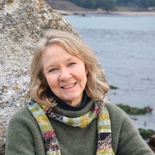 Meg Waite Clayton, a light-skinned woman with shoulder-length blonde hair, a grayish-green sweater, and a thin scarf, smiles at the viewer. She sits in front of a rock overlooking a body of water.