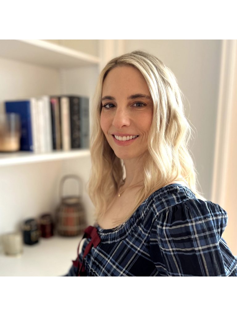 Mara Rutherford, a light-skinned woman with shoulder-length blonde hair, wearing a plaid shirt, smiles at the viewer. She is posed at a three-quarter angle beside a white bookcase.
