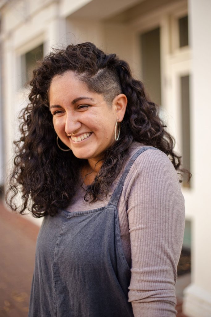 Andrea Beatriz Arango's body is tilted away from the camera, her gaze downward as she smiles widely. She has brown eyes, tan skin, a gold septum piercing, and dark, curly hair that is shaved close on one side. She is wearing a gold hoop earrings and a mauve, ribbed long-sleeve shirt with indigo-colored overalls over it.