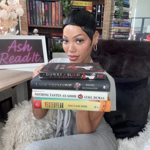 Bookstagrammer Ashley Lauren sitting in her book nook holding a stack of four books.
