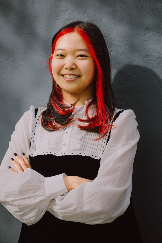 Author K-Ming Chang is standing against a gray wall, smiling to the camera with her arms crossed. She has tan skin, dark eyes, and black hair with red highlights. Her nails are painted black. She is wearing a long-sleeved, frilly white blouse with a black vest over the top and a silver chain necklace with a heart shaped pendant.